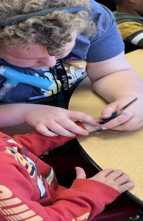 A 12th Grade student guides a preschool student’s fingers to hold a pair of scissors. 