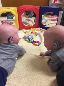 twin infants wearing hearing aids are playing with toys in front of mirrors