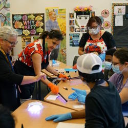 A group of students, a teacher and an interpreter watch as a woman shows them how to dry resin using a UV light.