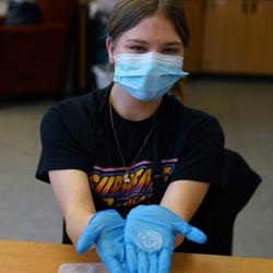  A student wearing a paper mask and gloves holds out a sparkly blue circle made of resin.