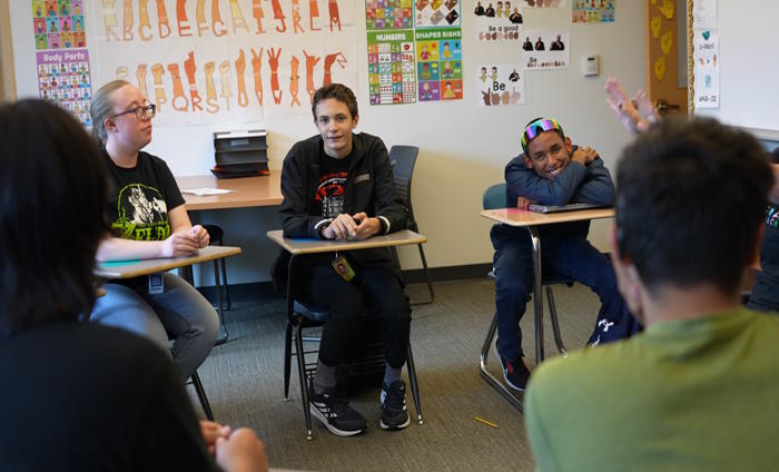 Three students sit in their desks facing the camera.  Two students sit facing the other students and back to the camera.