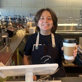 A Bridges to Life student smiles behind a counter with a tablet while holding a cup of coffee in a to-go cup. 