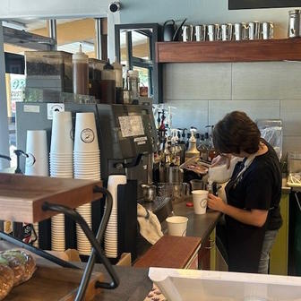 A student stands next to a variety of coffee preparation tools and pours milk into a container.