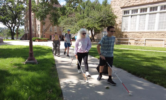 Three students from Adams and a teacher walk on a sidewalk using their canes.