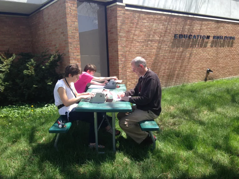 Two students and a teacher taking advantage of the sunny day, brailling outside at a picnic table.
