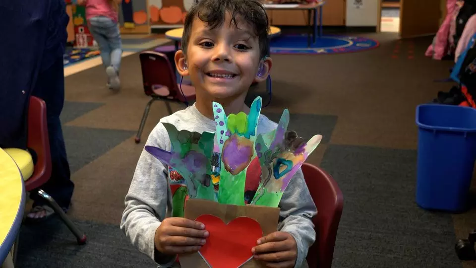 Young student, in the School for the Deaf, proudly holds up his heart art that has three "I Love You" handshapes coming out of the heart. Student grins happily.