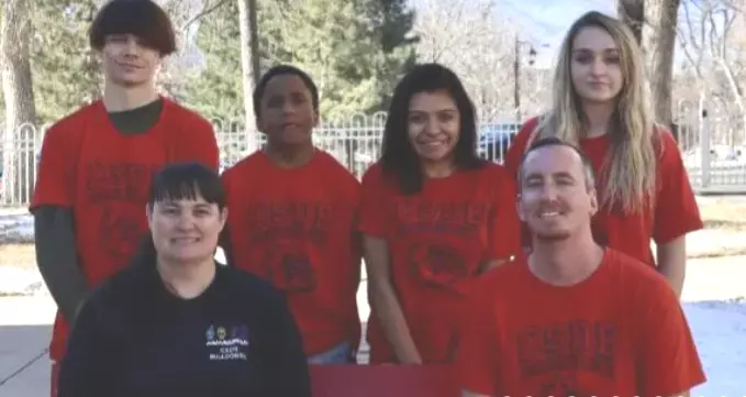 Four students with red shirts stand behind two teachers sitting down.