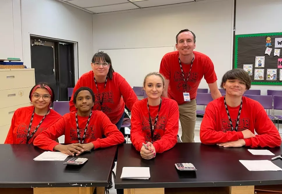 Four students in red shirt sit at a table and two teachers in red shirts stand behind them.