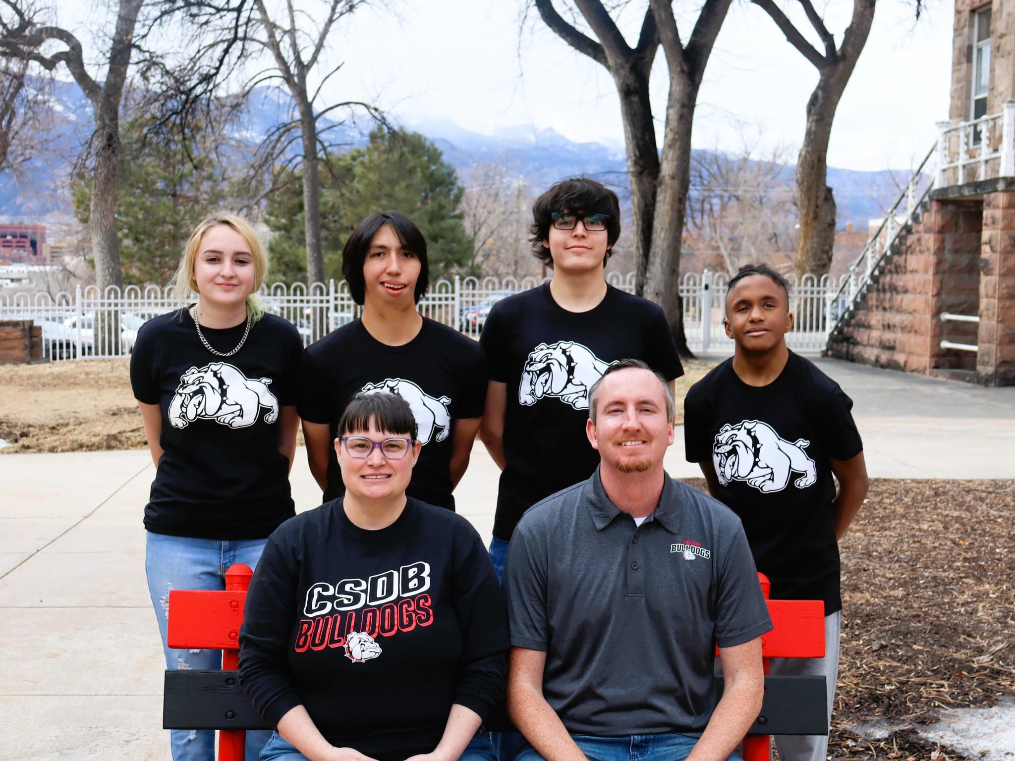  Four students in black bulldog shirts stand in the back, while two coaches sit in front of them, with mountains and trees in the background