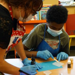  A student spreads resin in a mold using a wooden stir stick, while a teacher watches.