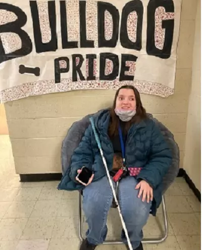 Cheyenne, holding a white cane, sits under a Bulldog PRIDE banner