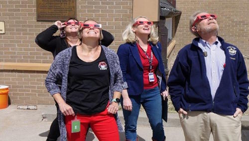 4 adults wearing eclipse glasses look at the eclipse.