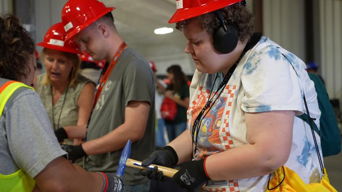 Two students and a teacher, wearing red hard hats are learning about construction tools.