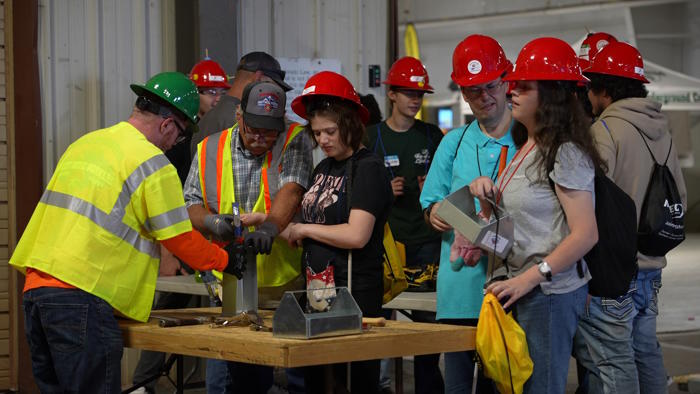 A group of students and teachers, wearing red hard hats, watch a demonstration on how to use a construction tool.