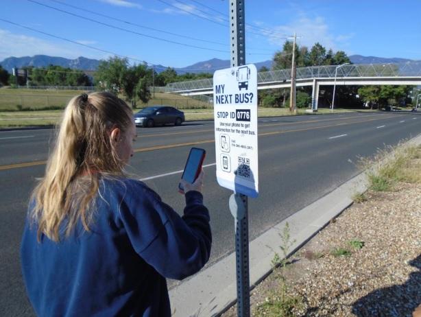 DHH BtL student using her phone to get city bus information from a bus stop sign during a bus training by a Travel Trainer from Mountain Metro Transit.