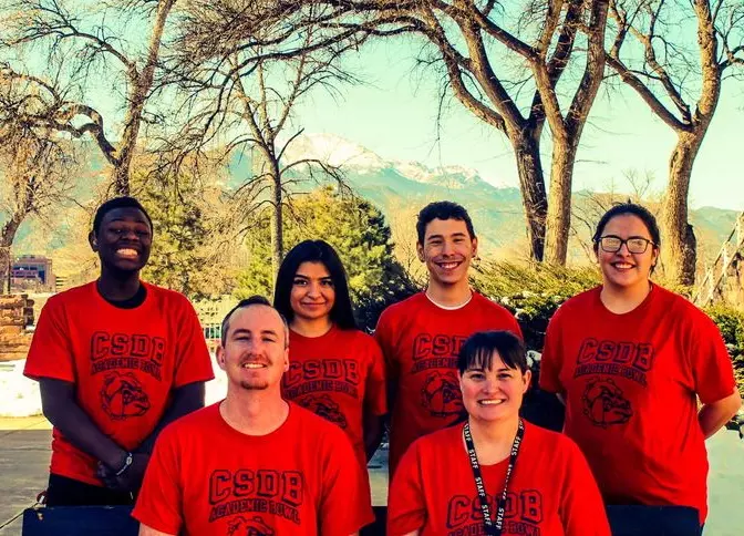 left to right: Gideon (12th grade), Mariah (11th grade), Ethan (12th grade), Marie (12th grade) and in front, coaches, Max and Amy.  The group is wearing matching red bulldogs shirts, smiling at the camera with mountains in the background