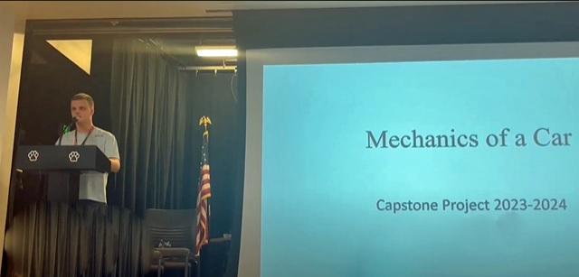 Male student presents behind a podium and next to the screen "Mechanics of a Car"