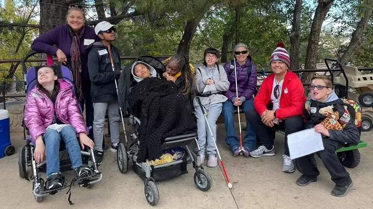 5 students with canes and wheelchairs and four adults smile at the camera while outside at the zo