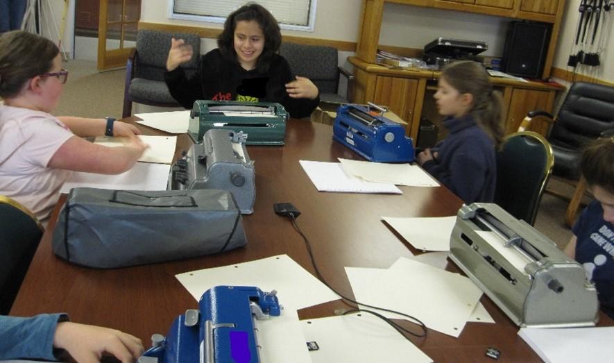 Alt text: 5 contestants sit with their braillers at a long table during the 2023 Denver Metro Braille Challenge Contest.