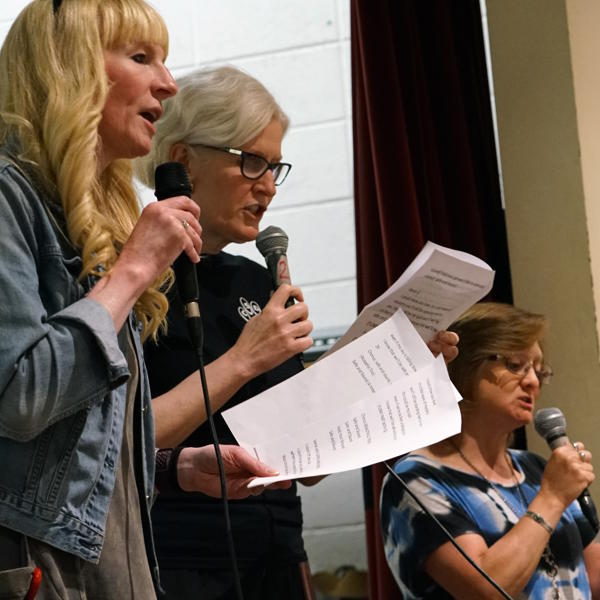 Three teachers stand on the stage, holding music sheets and microphones.  They are singing into the microphone.