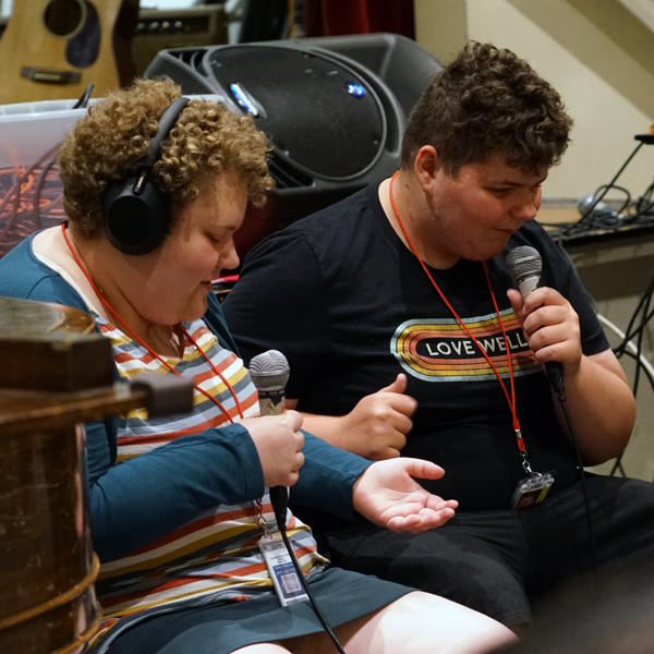 Two students sit in chairs. They are holding and singing into a microphone.