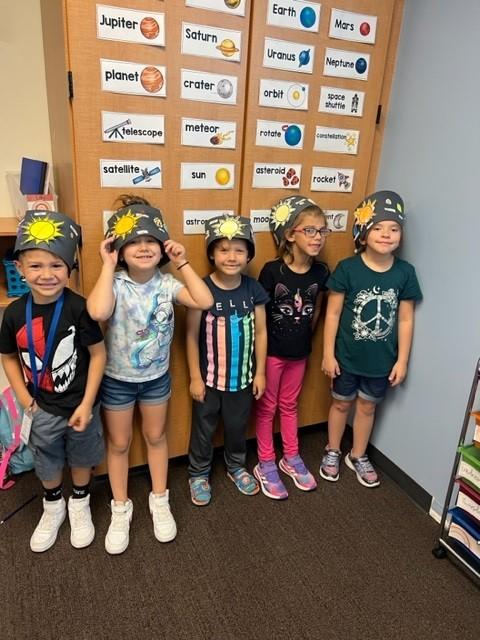 Five young students pose wearing space hats and smile in class