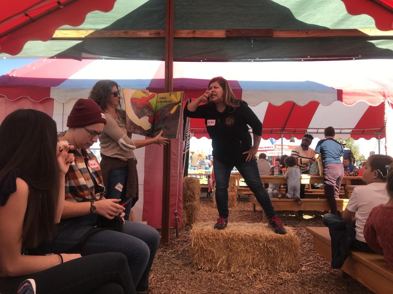 A woman ASL storyteller at work on a haybale in a barn while others watch