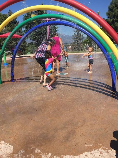 Three children and two ladies duck under four multicolored sprinklers in the park