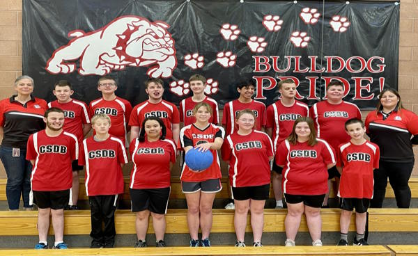 16 people, goalball players and coaches stand in the bleachers in front of a Bulldog Pride banner.