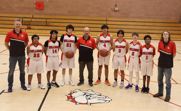 10 people, student basketball players and coaches, stand on the CSDB gym floor behind the Bulldog logo.