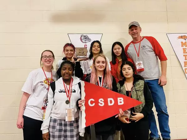 eam of 6 high school girls and 2 coaches stand for the photo with CSDB red Pennant banner. One girl holds the trophy.