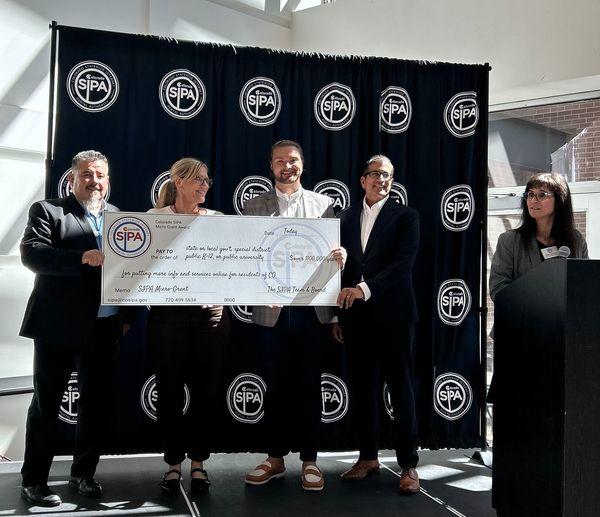 Three men and a woman hold an extra-large check and smile, while another woman stands at a podium next to them.