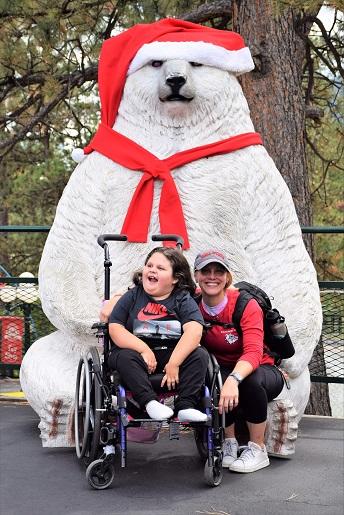 Teacher and student, in a wheelchair, post for a photo in front of a giant polar bear statue, wearing a Santa red hat and scarf
