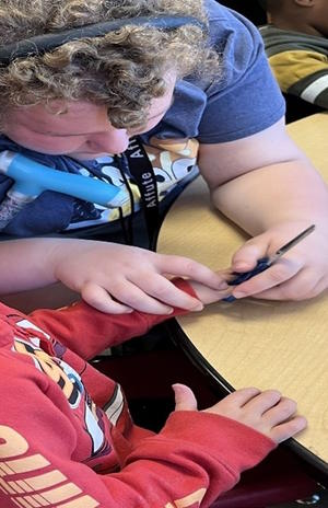 A students guides a preschool student's fingers to hold a pair of scissors.