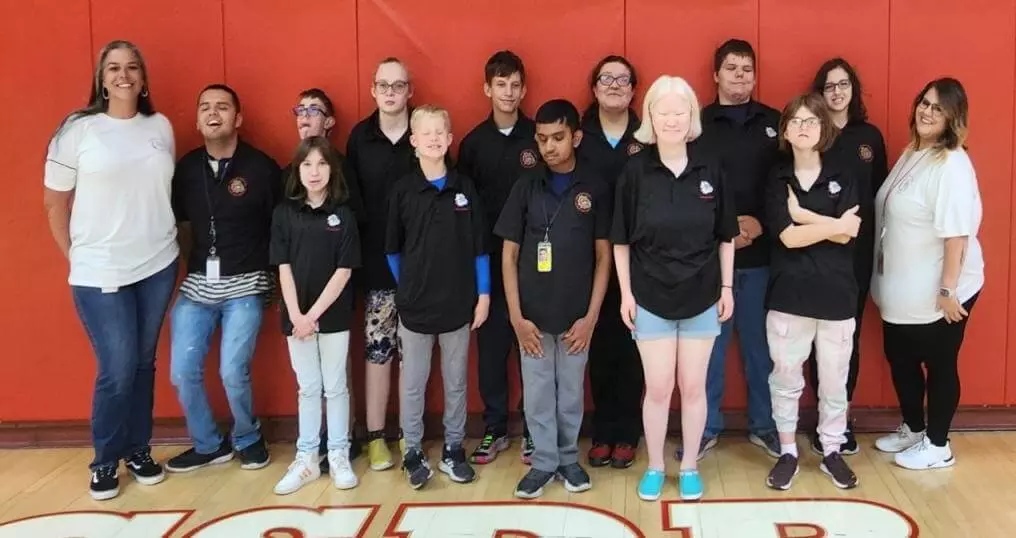 12 members of the Special Olympic bowling team (wearing black shirts) and 2 coaches stand in front of the red mat wall.  