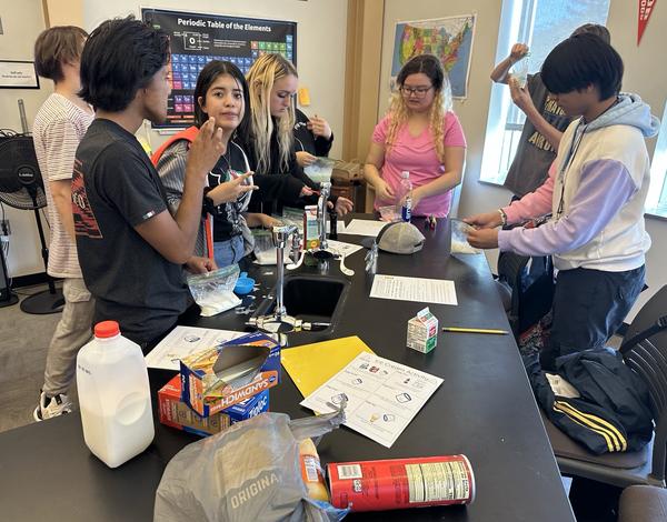Seven students perform a group experiment around a science lab table.