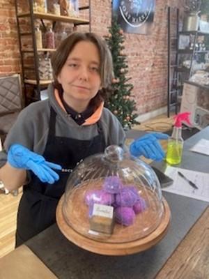 Caption:  Female DHH BtL student is standing behind a display of bath bombs that she assembled for her work study site: Rocky Mountain Soap Factory 