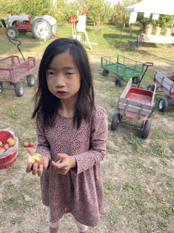 A student looks at the camera and is holding an apple.  There are wagons in the background.