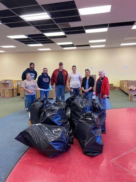(Pictured is 2 CSDB employees and 6 Toys for Tots volunteers and coordinators standing in a warehouse of toys with 10 large black bags full of donated items)