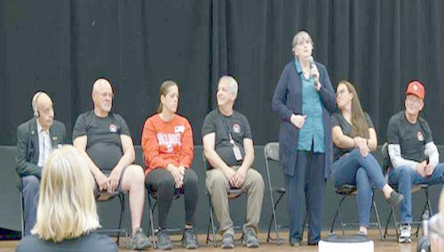 6 alumni sit on the stage, 1 alumni is standing and talking into a microphone