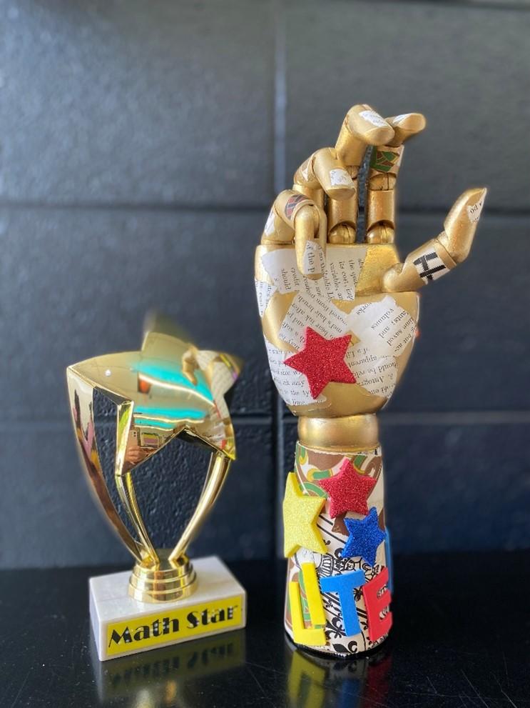 Two trophies: left, rounded gold star with marble base- text ‘Math Star’; right, wooden hand with flexible fingers spray painted gold with random modge-podged text, foam star stickers and 3D letters that spell out ‘LITERACY.’