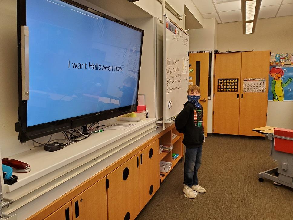 Boy stands in classroom next to large monitor that reads, "I want Halloween now."