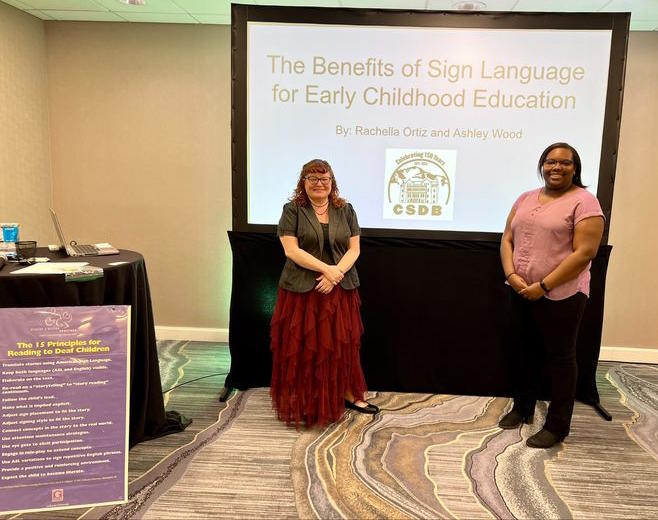 wo ladies stand in front of a large screen containing text "The Benefits of Sign Language for Early Childhood Education."