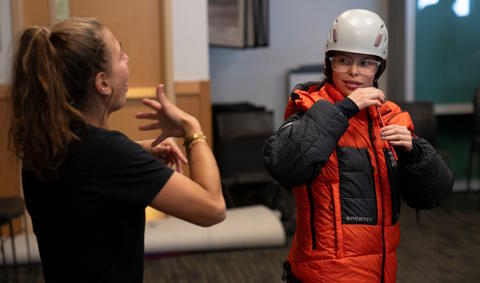 A student wears a heavy coat and a helmet, while Shayna communicates with her in ASL