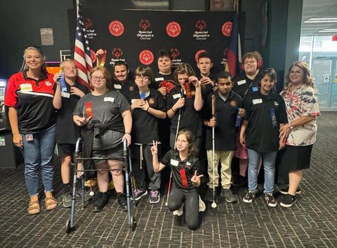 A group photo of 11 smiling students and two staff members in front of a large Special Olympics of Colorado backdrop. Every student holds a ribbon or has on attached to their shirt.