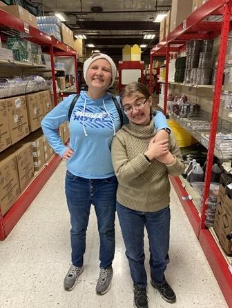 Two female BtL students pose for the photo in a warehouse setting, while stocking shelves at Envision Xpress