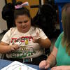 A student holds a sticker and smiles, while a student aide watches.